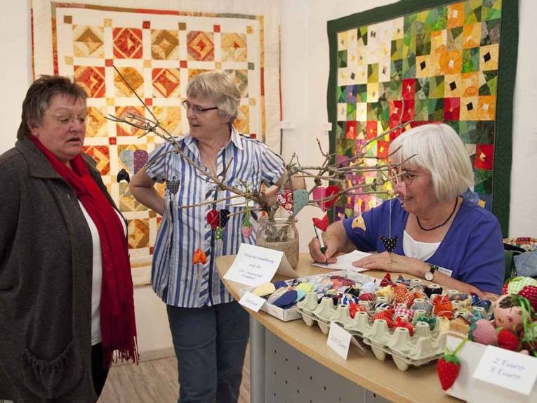 Drei Frauen stehen an einem Tisch mit Handarbeitsutensilien, im Hintergrund hängen Patchwork-Decken an den Wänden.