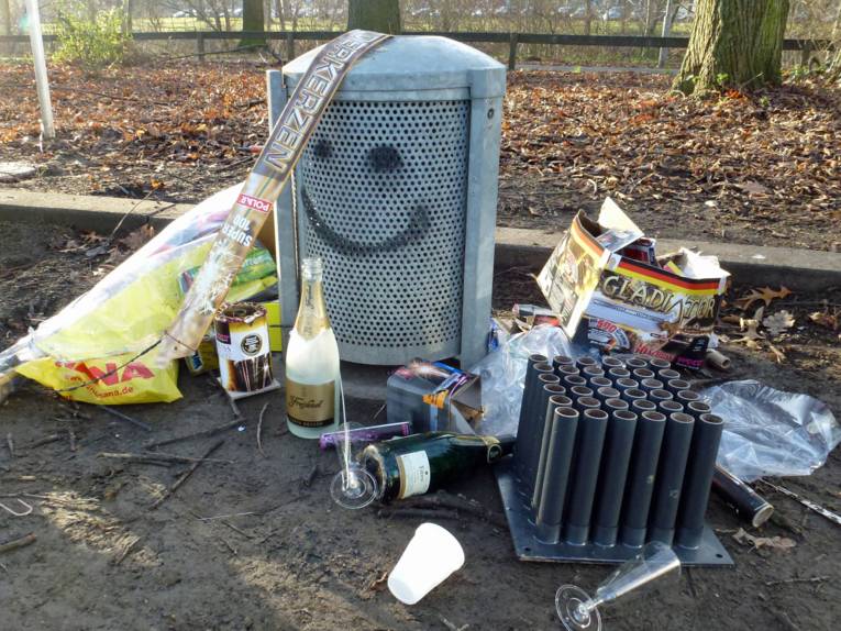 Leere Sektflasche, Plastikgläser und abgebrannte Feuerwerksartikel um einen Mülleimer herum.