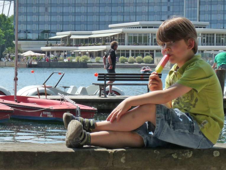 Junge sitzt an einem See auf einer Ufermauer und lutscht an einem Eis.