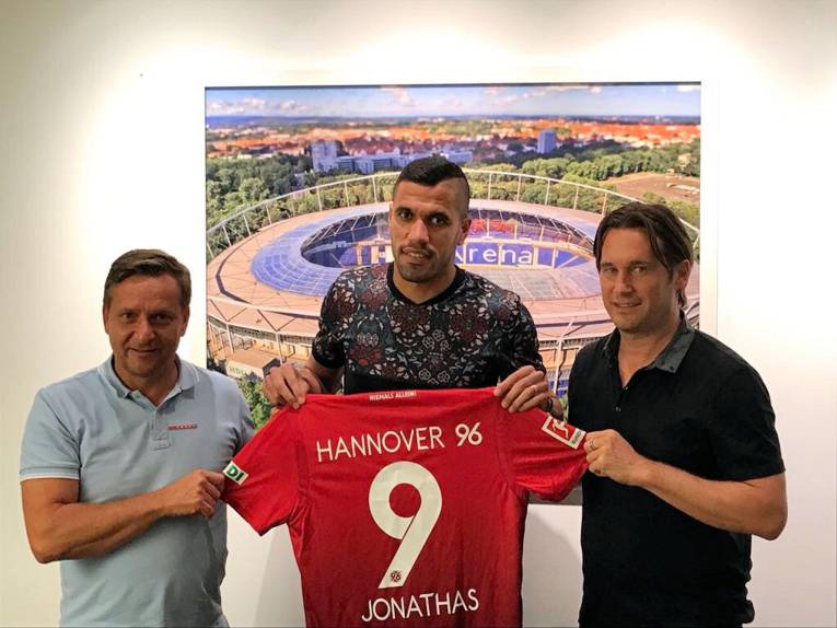 Drei Männer halten ein Trikot von Hannover 96 mit der Rückennummer 9 und der Aufschrift Jonathas.