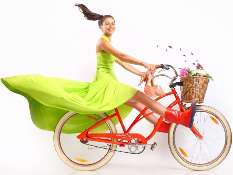 Foto: Junge Frau im grünen Kleid und Gummistiefeln auf einem Fahrrad, in der Hand ein Paar rosafarbene High Heels.