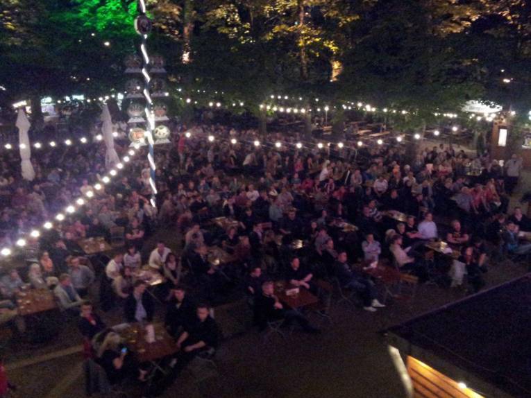Viele Menschen in einem Biergarten, die auf eine Leinwand schauen.