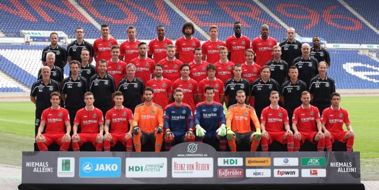 Spieler, Trainer und Betreuer einer Fußballmannschaft in vier Reihen hintereinander in einem Stadion.