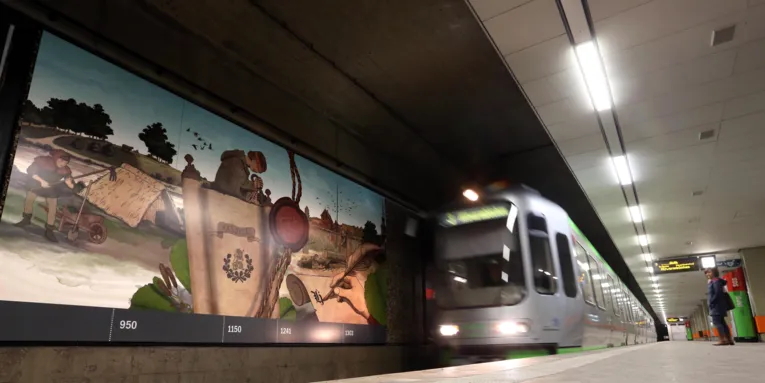 Eine Bild-College an der Wand eines Stadtbahn-Gleises und eine einfahrende Stadtbahn.