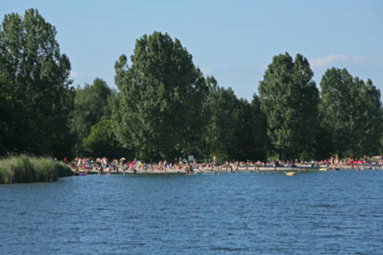blaues Wasser, im Hintergrund badende Menschen an der Uferzone des Altwarmbüchener Sees