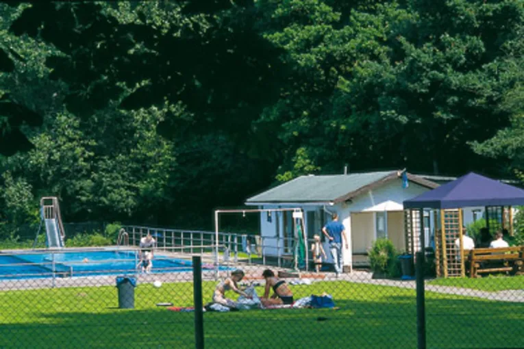 Liegewiese mit zwei Badegästen, im Hintergrund ein Schwimmbecken mit Rutsche, rechts ein Gebäude und ein blauer Gartenpavillion