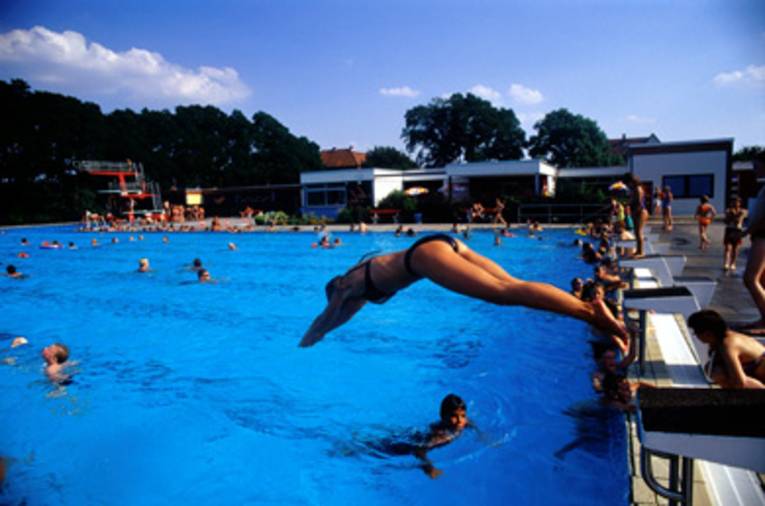 Eine Frau beim Kopfsprung vom Startblock ins Schwimmbecken, im Wasser schwimmende Menschen, im Hintergrund ein roter Sprungturm und weisse Flachdachgebäude