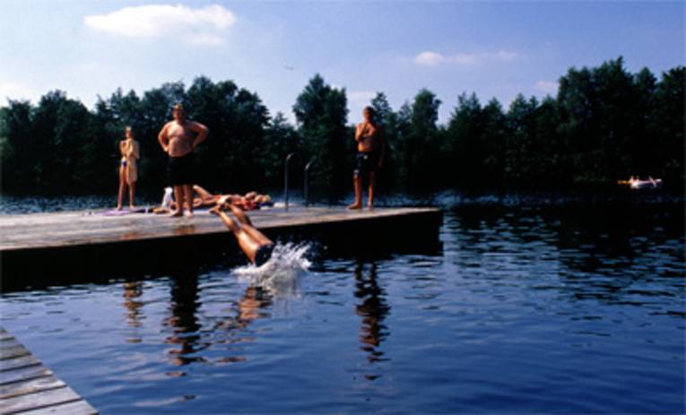 ein Holzbadesteg auf dem Kinder stehen, ein Kind springt gerade mit einem Kopfsprung ins Wasser, im Hintergrund ein Paddelboot