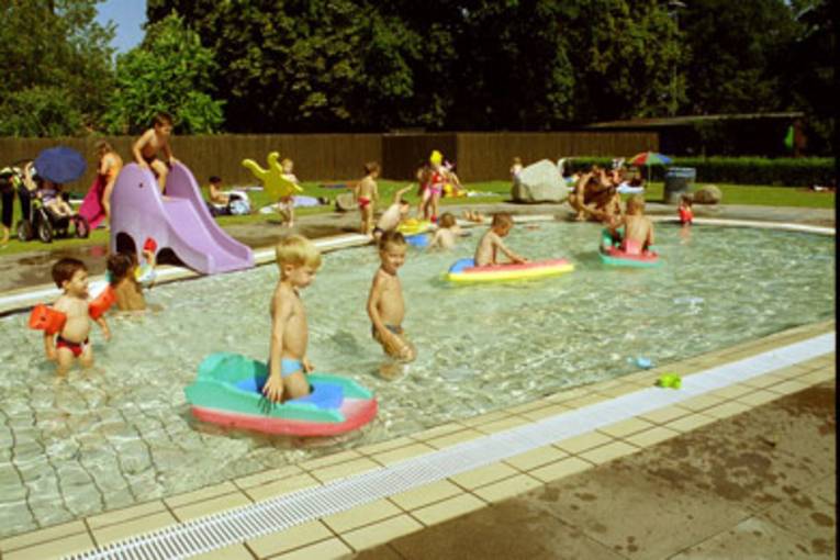Kleine Kinder toben mit Wasserspielzeug und auf einer Rutsche in einem Nichtschwimmerbecken unter freien Himmel.