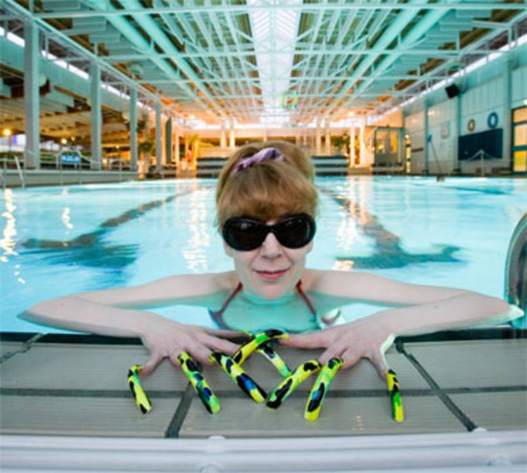 Eine Frau mit großer Sonnenbrille und sehr langen, bunt lackierten Nägeln lehnt sich an den Beckenrand vom Schwimmerbecken.