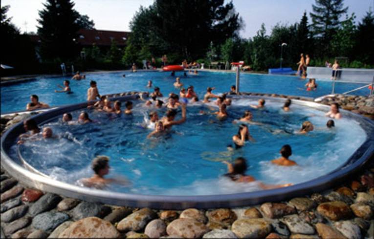 ein rundes Schwimmbecken, in dem sich Menschen vergnügen, im Randbereich ist eine starke Wasserströmung zu erkennen, das Becken grenzt an ein normales Schwimmbecken an