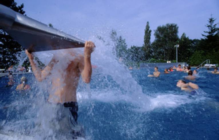 Ein Junge hat sich am Kopf der Wasserschwallbrause hochgezogen, der Körper wird vom Wasser umströmt, im Hintergrund im Wasser spielende Kinder
