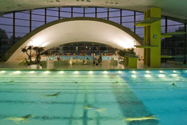 einige Badende tauchen im beleuchteten Schwimmerbecken, im Hintergrund eine Fensterfront