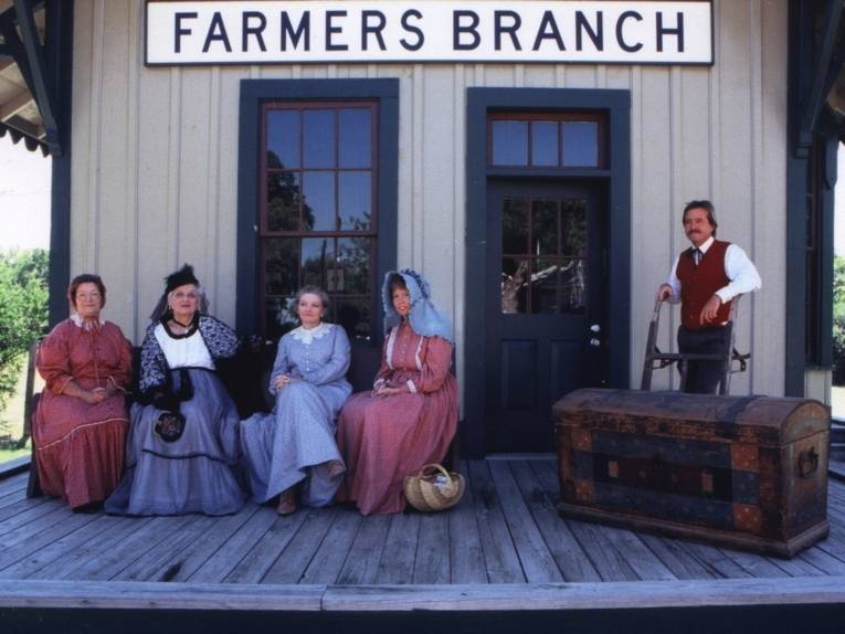 Vier Frauen in Trachten sitzen auf einer Bank vor einem Häuschen mit der Aufschrift Farmers Branch.
