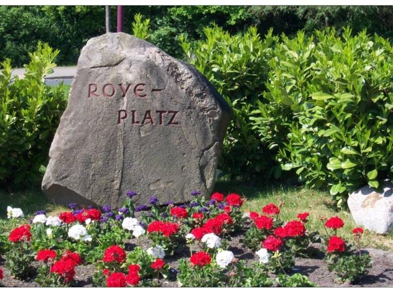 Ein großer Steine mit der Gravur Roye-Platz umgeben von Sträuchern und Blumen