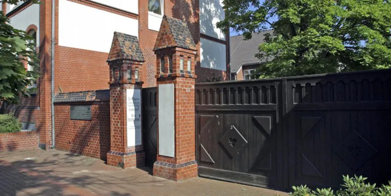 Vor dem Eingang zur Gedenkstätte Ahlem. Teilweise Steinmauer und der Eingang ist zwischen zwei genauerten Säulen, die aussehen wie kleine Türme.