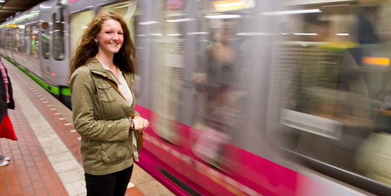 Junge Frau steht in einer U-Bahnstation und eine Bahn fährt ein.