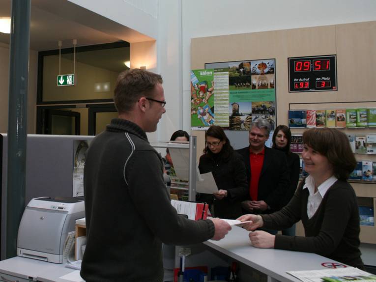Eine Frau an einem Informationstresen, die einem Mitarbeiten ein Dokument überreicht. Dahinter stehen noch weitere Personen in der Schlange. Im Hintergrund sind außerdem Broschürenständer zu sehen.