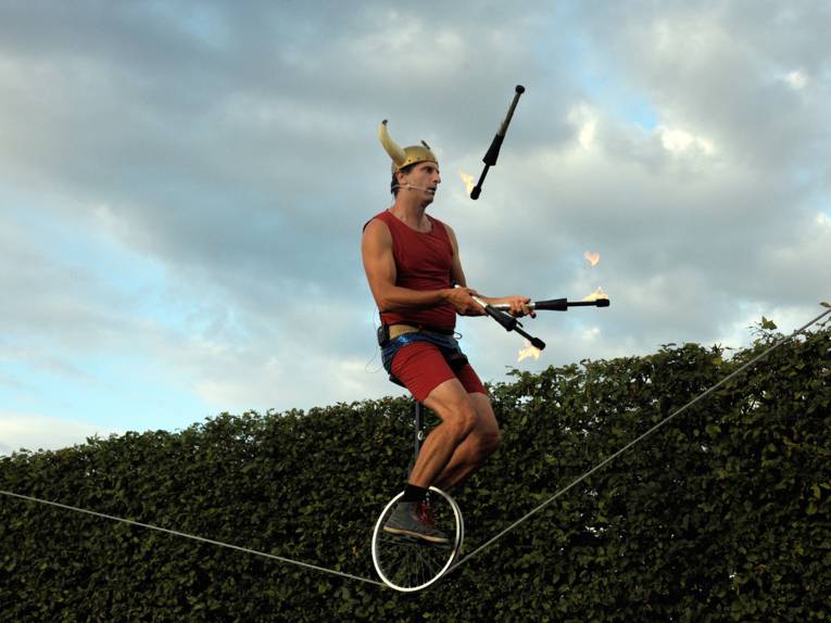 Ein Mann führt mit einem Einrad auf einem Seil in einigen Metern Höhe und jongliert brennende Kegel