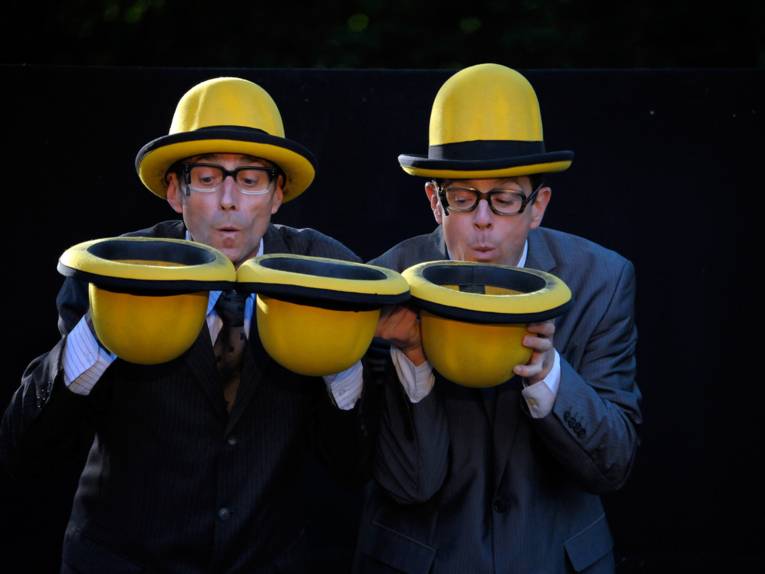 Zwei Männer tragen jeweils einen gelben Melonenhut auf dem Kopf, einer hält zwei weitere Hüte, der andere einen weiteren Hut in den Händen