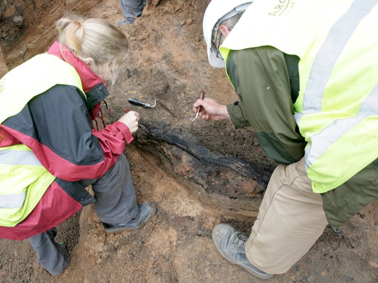Zwei Mitarbeiter von ArchaeoFirm legen ein verbranntes Holzfundament frei