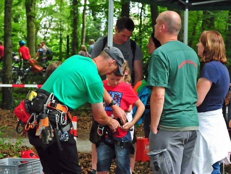 Junge wird vor dem Baumklettern von Mitarbeitern der Waldstation mit Gurten gesichert