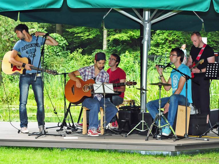 Eine fünfköpfige Band spielt auf einer Bühne, die auf einem Wiesenstück im Wald aufgebaut ist.
