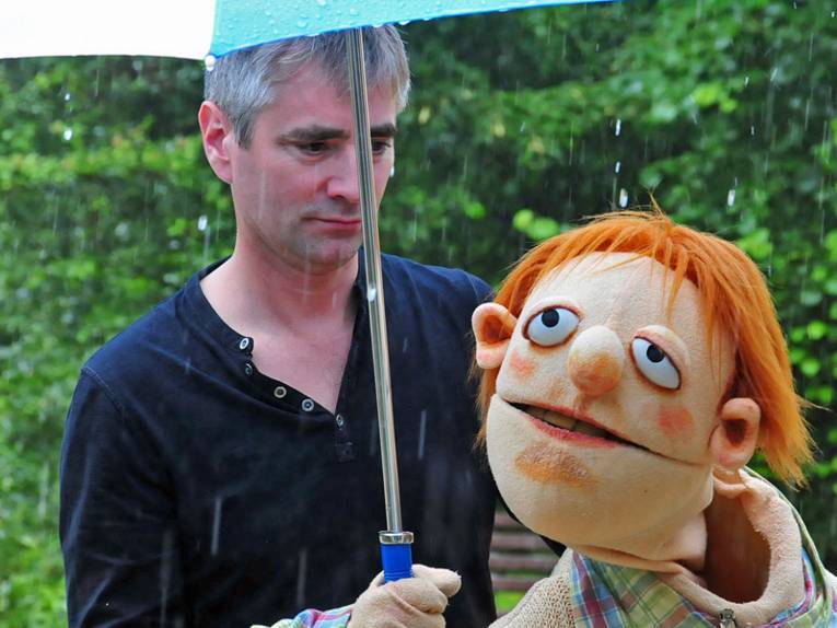Ein Schauspieler agiert bei Regen mit einer Handpuppe unter einem Regenschirm