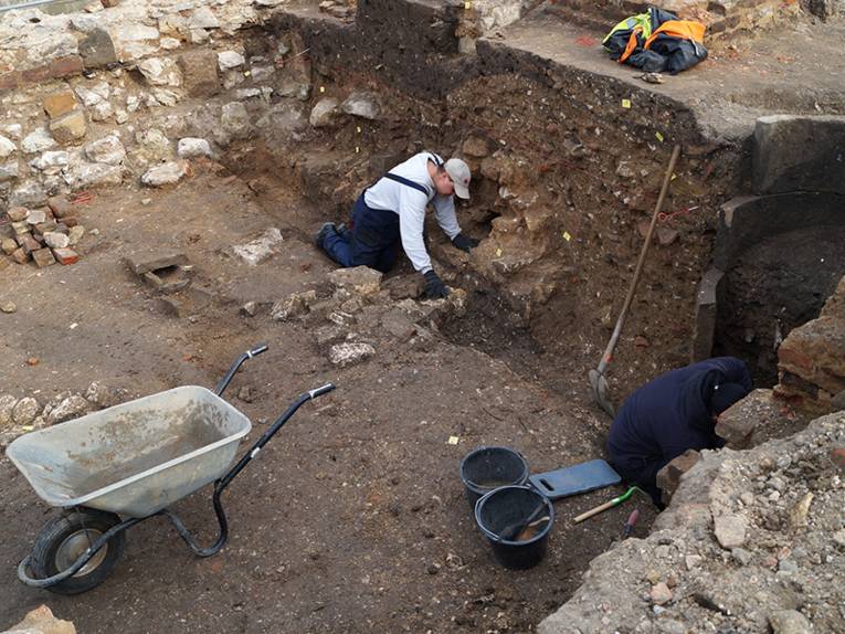 Zwei Arbeiter schachten das Grabungsfeld aus. Links steht eine Schubkarre, rechts die Reste eines Brunnens.