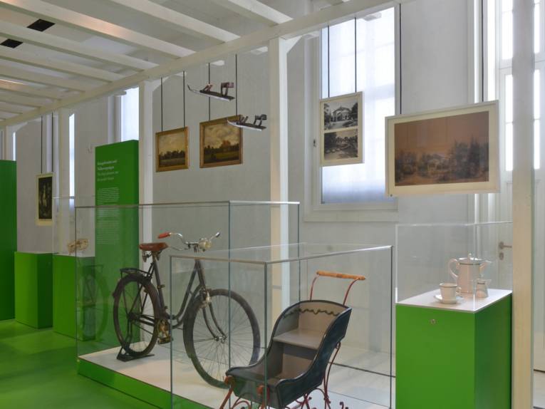 Ein Blick in den Gartenflügel im Museum Schloss Herrenhausen: Ein altes Fahrrad und ein alter Kinderwagen in Glasvitrinen, im Hintergrund diverse Bilder