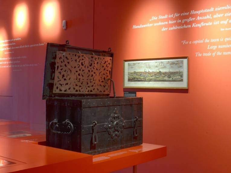 Eine massive Kiste, in der in früheren Zeiten Geld aufbewahrt wurde, inmitten des roten Barockflügels im Museum Schloss Herrenhausen; an der Wand im Hintergrund hängt ein Stich, der Hannover im Mittelalter zeigt