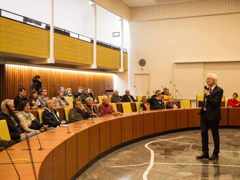 Bernd Strauch mit einem Mikrofon im Ratssaal, auf den Plätzen gut zwei Dutzend Gäste