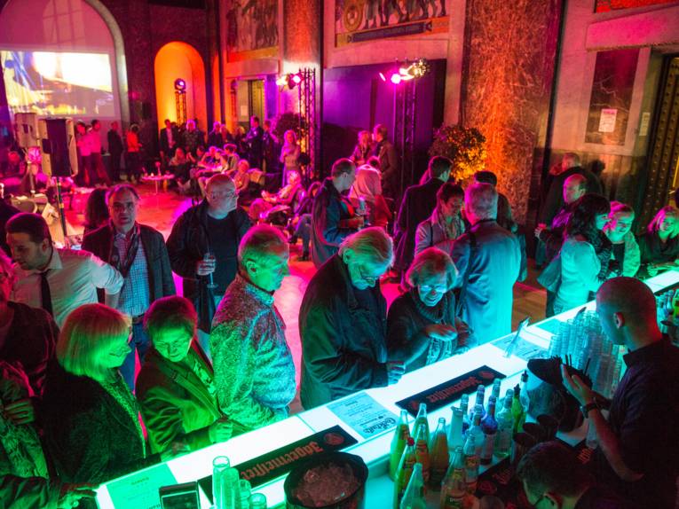 Viele Personen stehen an der Bar im Mosaiksaal und sind in ein türkis-rotes Licht getaucht
