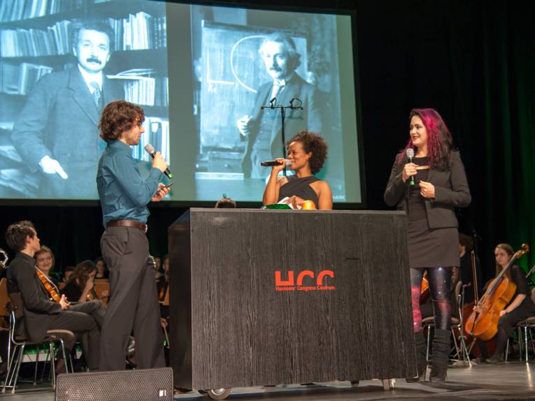 Ein Mann und zwei Frauen stehen auf einer Bühne, alle halten Mikrofone. Im Hintergrund sind Musikerinnen und Musiker eines Orchesters, auf der Leinwand sind zwei Fotografien bedeutender Wissenschaftler.
