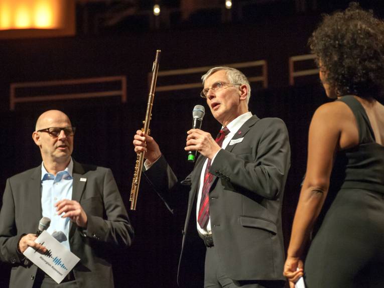 Zwei Männer und eine Frau stehen auf einer Bühne, der Mann in der Mitte spricht in ein Mikrofon und hält eine Querflöte in der anderen Hand.