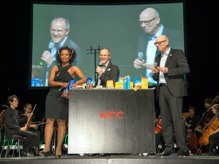 Eine Frau und zwei Männer stehen auf einer Bühne, auf einem Tresen zwischen ihnen liegen Kekse und anderes Gebäck.