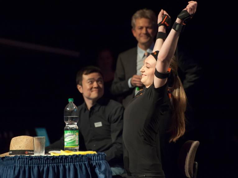 Eine Sportlerin streckt sich auf der Bühne, auf dem Tisch vor ihr sind ein Strohhut, ein Wasserglas und eine Wasserflasche.