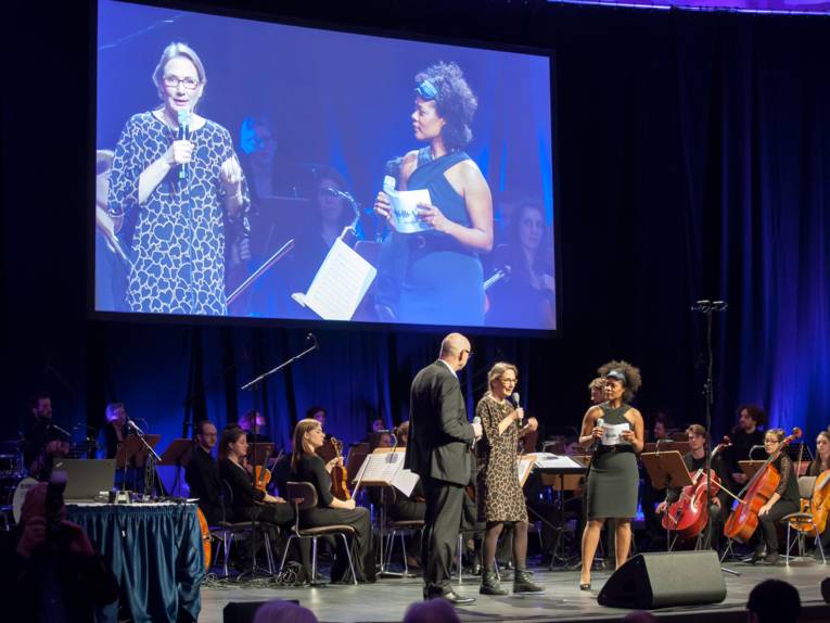Blick auf die Bühne im Kuppelsaal, die Leinwand zeigt eine Live-Aufnahme des Bühnenprogramms.