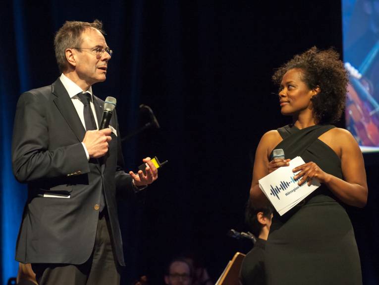 Ein Mann (links) und eine Frau stehen auf einer Bühne, beide haben ein Mikrofon in der Hand. Die Frau hält außerdem weiße Moderationskarten mit Logo der Hörregion Hannover.