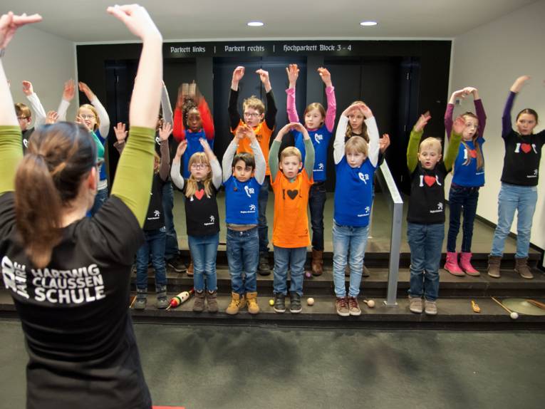 Eine Frau steht vor einer Gruppe von Kindern. Sie macht etwas vor, die Kinder machen es nach.
