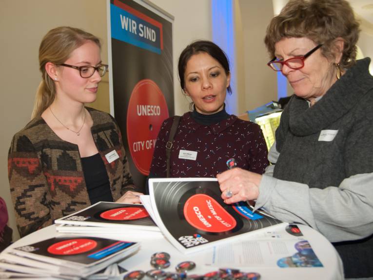 Drei Frauen stehen an einem Stehtisch und blättern durch eine Broschüre.