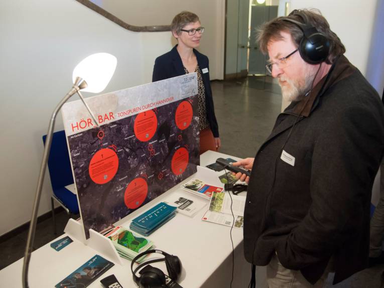 Infostand mit Ausstellungsstücken, davor steht ein Mann und trägt Kopfhörer.