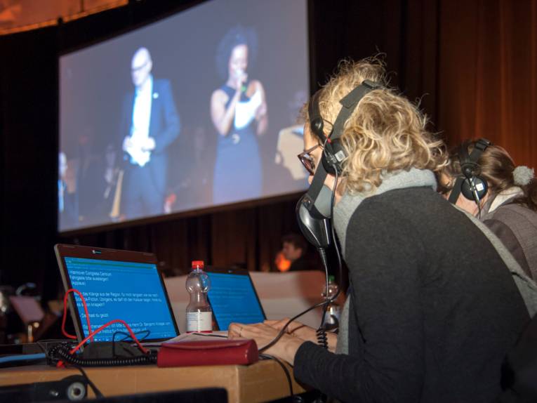 Zwei Frauen sitzen an Laptops und tragen Kopfhörer und aussenschallisolierte Mikrofone vor dem Mund.
