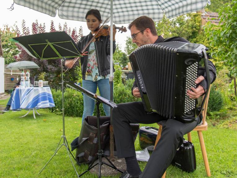 Eine Geigenspielerin und ein Akkordeonspieler unter einem Sonnenschirm im Garten.