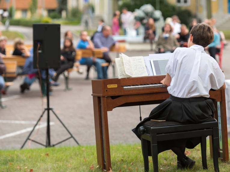 Eine Frau spielt draußen auf einer Rasenfläche Klavier. Mehrere Zuschauer sitzen davor.