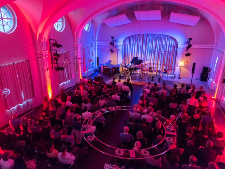 Ein Saal mit Publikum von oben fotografiert. Auf der Bühne spielt ein Mann Klavier.