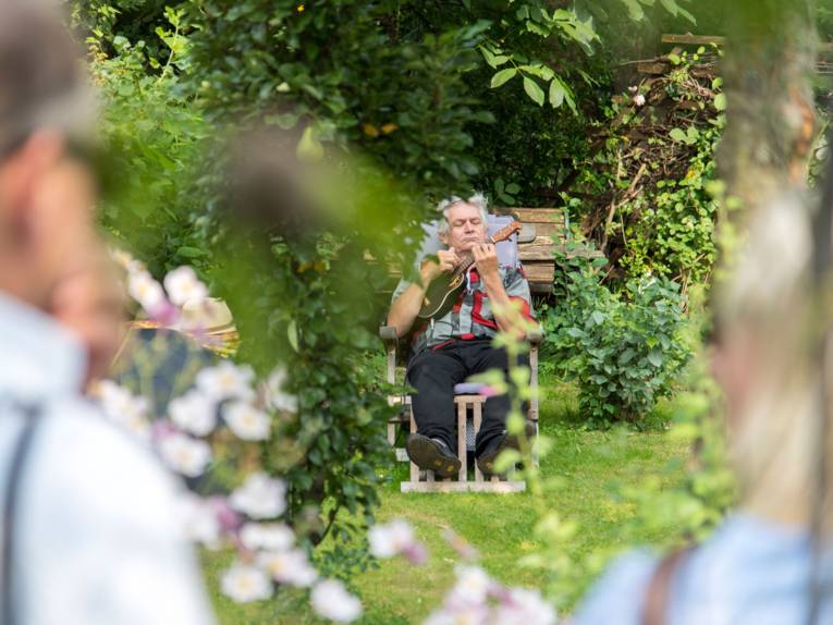 Ein Mann mit Mini-Gitarre auf einem Stuhl im Garten. Vorne im Bild sind Zuhörer zu sehen.