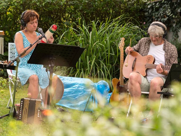 Eine Frau und ein Mann sitzen in einem Garten. Beide tragen ein Kopfhörer und spielen ein Instrument. sie haben weitere Instrumente dabei.