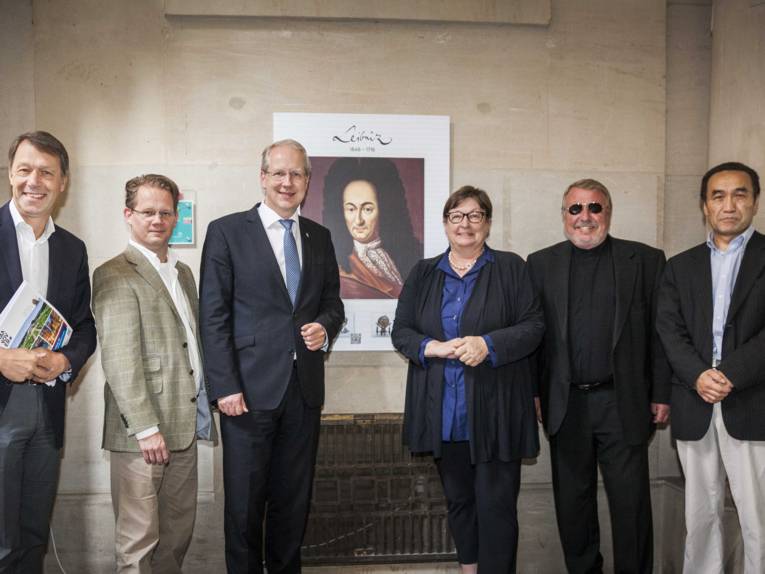 Fünf Männer und eine Frau vor einer Bildtafel, die an einer Wand hängt.