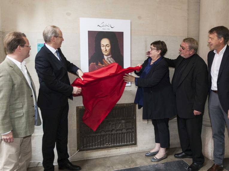 Vier Männer und eine Frau nehmen ein rotes Tuch von einer an der Wand hängenden Bildtafel herunter.